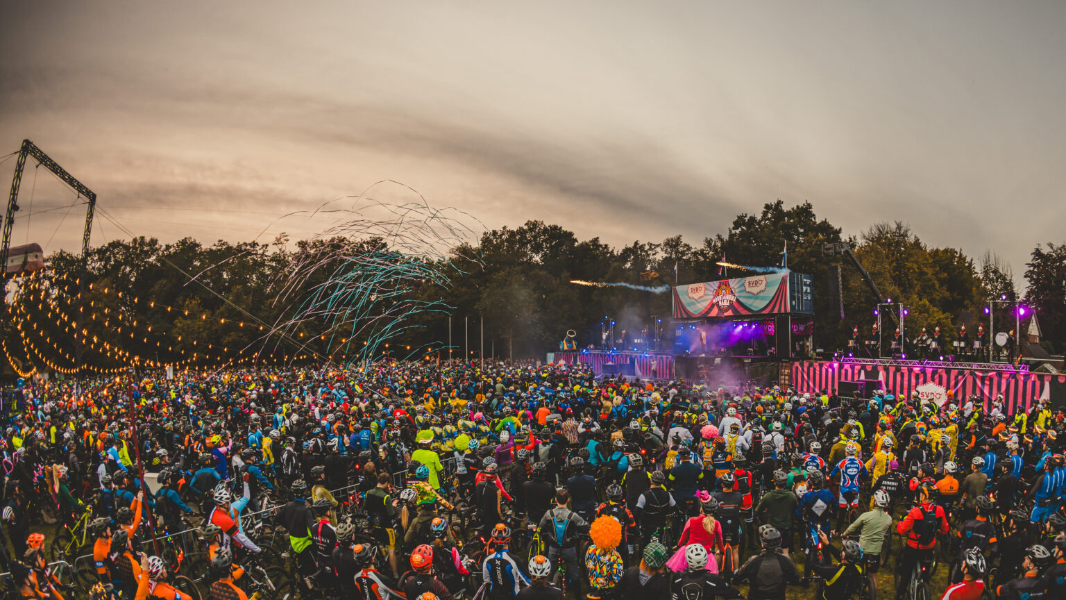 Groot outdoor evenement met duizenden deelnemers op mountainbikes, kleurrijke verlichting, confetti en een festivalpodium tijdens de startshow.